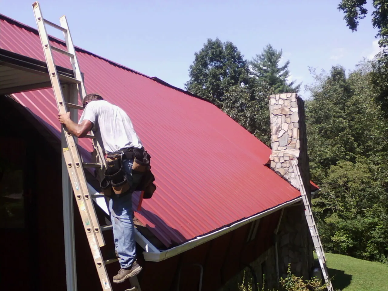 Roofer on a ladder beside a red metal roof and stone chimney — Soffit & Fascia Repair by Roofing Brothers in North Hills